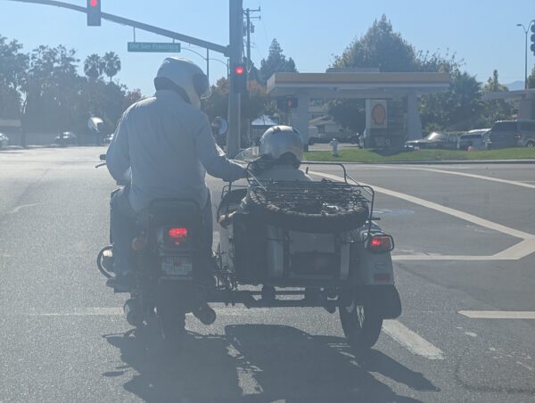 A motorcycle with a side car waits at a red light at Old San Francisco Road. The pilot has turned towards the smaller passenger in the side car, and has their hand on the passenger's shoulder.