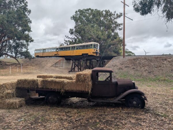 A vintage yellow Key System "bridge unit" crosses a short wooden trestle at the Western Railway Museum. In the foreground, an even older, thoroughly rusted flatbed truck, with straw bales stacked on the bed, sits in leaf-covered mud.