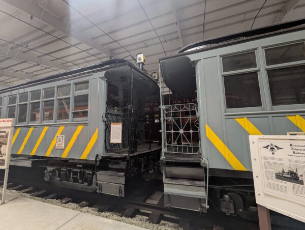 Two long-serving passenger rail cars, painted grey with yellow stripes, sit in restored condition at the Western Railway Museum.