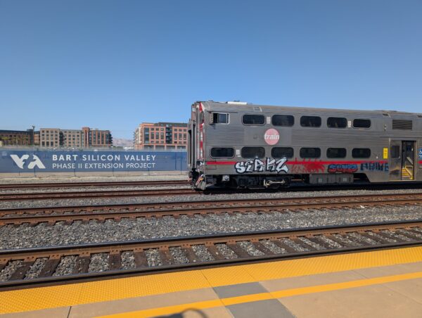 A view from Santa Clara Caltrain. On the left is a fence with a sign that reads "BART SILICON VALLEY PHASE II EXTENSION PROJECT" and on the right is a Caltrain gallery car with some graffiti on the lower level.