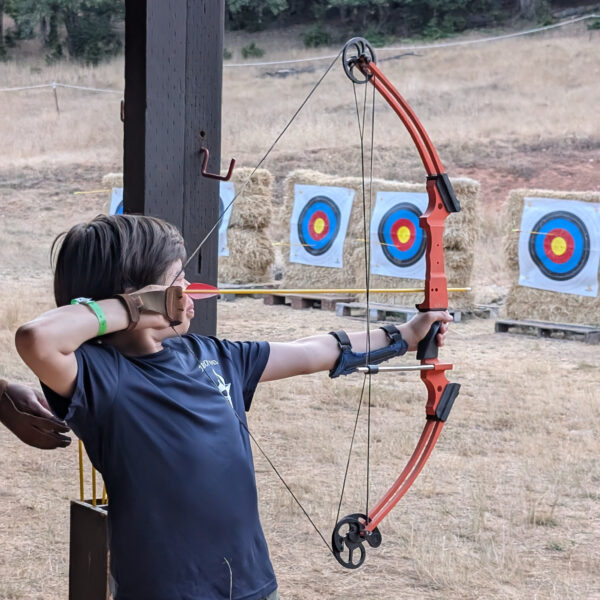 A scout in a blue shirt pulls back on the string of a bow, preparing to fire at a target affixed to a hay bale.
