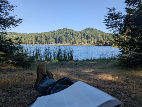 A view of mountains beyond a lake, near the shore. Danny's feet and legs are visibly stretched out, with a magazine on the lap.