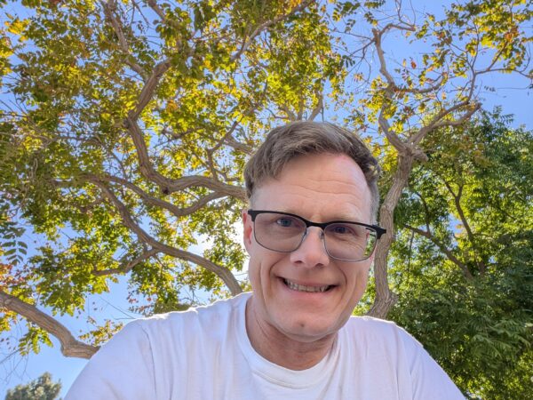 Danny, wearing new glasses and a white t-shirt, smiling. Behind him are the casually twisting branches of a tree beneath a sunlit blue sky.