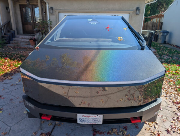 A Cybertruck, viewed from the front, with a shiny rainbow effect in the surface treatment. Three colorful maple leaves rest on the expansive windshield.