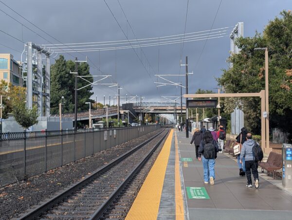 A view down the tracks at the Sunnyvale Caltrain station. The overhead wires are framed by dark clouds breaking in the distance.