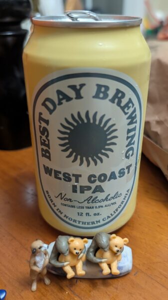 A can of "Best Day Brewing West Coast IPA Non-Alcoholic" and just in front of it sit small plastic figurines: two sloths holding teddy bears as a meerkat looks on.