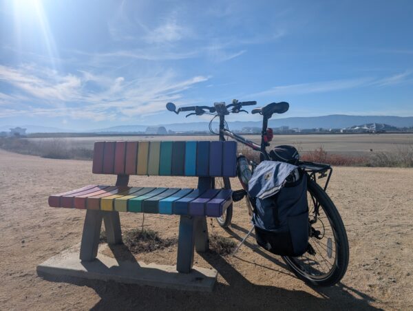 A bicycle leans against a bench. The bench is painted in a rainbow. The scene is on a dirt trail in the San Francisco Bay, a gleam of sunlight on the left and buildings in the distance, including a blimp hanger at Moffett Field, and a Google office.