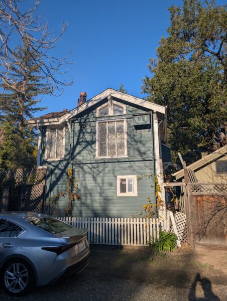 A an old wood two-story house as seen across a backyard fence. It has ornate windows.