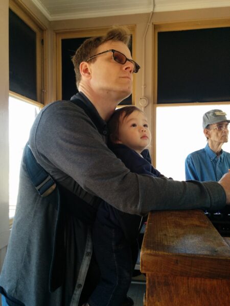 Danny leaning against the interlocking machine, with a baby strapped to his chest. Both he and the baby are looking up, together.