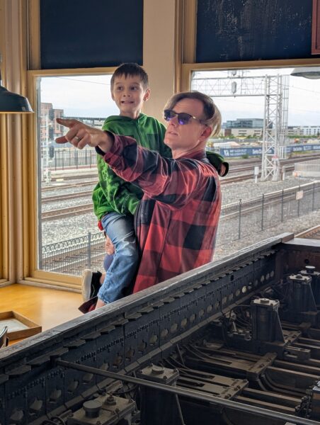Standing in front of the interlocking machine, Danny is holding a boy in his arm, pointing into the distance with his left hand. The boy is looking in the same direction.
