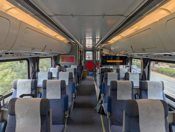 Within the upper level of a passenger train car. There is a red flag at the end of the aisle, and a view out the window of the tracks ahead.