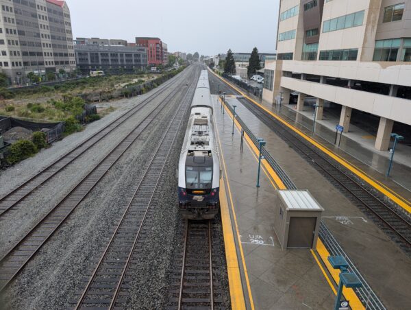 A five-car passenger train at center. The engine displays the word "Sacramento." There are three more tracks to the left and to the right, an island platform and one more track. The day is overcast, and a headlight can be seen in the distance.