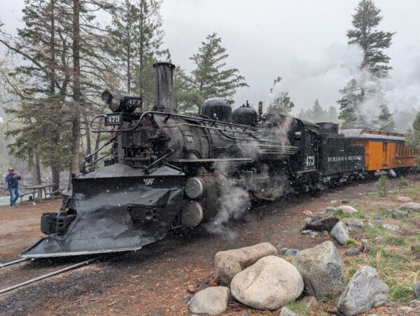 A steam engine, viewed from an angle toward the front, puffs out a bit of steam in the snow in the woods.