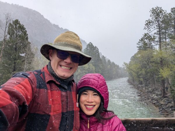 Danny and his Wife smile at the camera. Grey sky above, river below, snow blowing past.
