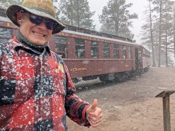 Danny poses for a selfie in front of D&S passenger car 350, "Alamosa" in the snow. He has snow sticking across his red flannel shirt, a thumbs up and a big smile.