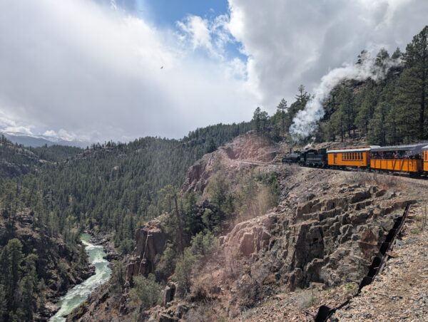 Upper right, a steam engine pulling wooden orange passenger cars. Lower left, the river. In the near ground, a rocky cliff, forest in the far ground.