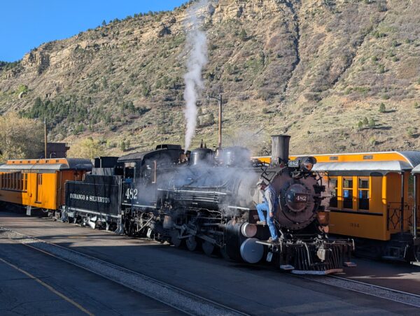 D&S 482 is in the Durango yard. The engineer is standing on the walkway along the boiler. The engine is shooting some steam straight into the sky. Mountains are visible as the backdrop.