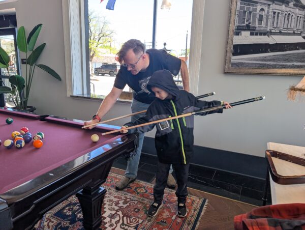 Danny and his younger son share the same pose, as Danny shows his son how to hold the pool cue to hit the ball.
