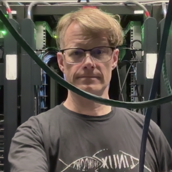 Danny in the datacenter, wearing a "Linux fish eats Windows fish" tee shirt. A network cable dangles under his chin in the foreground.